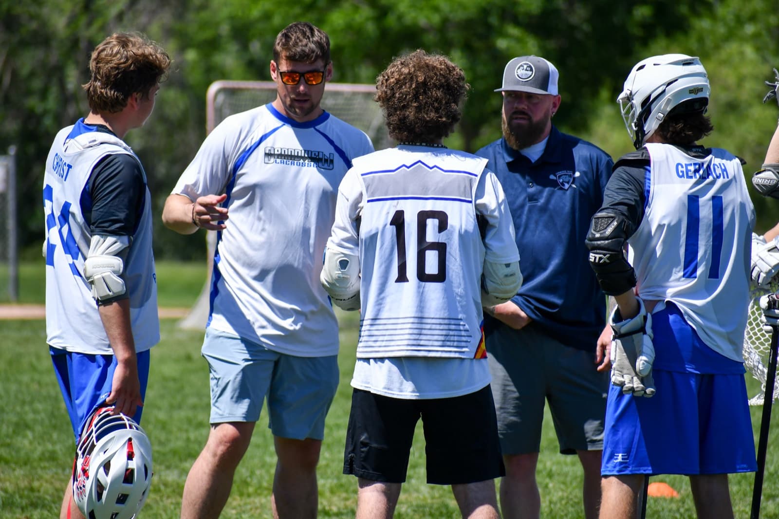 Two coaches talking with three players in a circle on a grass field, a lacrosse goal behind them.
